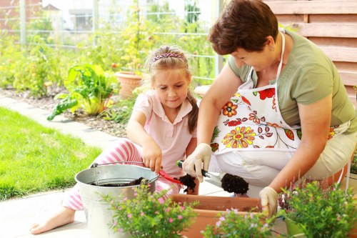 A woman and a young girl are working together in a well-maintained garden, planting and tending to flower pots placed on a paved patio area next to a garden shed. The woman, dressed in casual clothing and an apron, is guiding the girl as she carefully handles a small gardening tool, emphasizing outdoor activity and gardening care. Surrounding the pair are vibrant flowering plants and lush green foliage, with a tidy lawn visible in the foreground. In the background, a variety of garden elements such as potted plants, trees, and a wooden fence create a peaceful outdoor setting typical of residential gardens in Sanderstead. The scene is illuminated by natural daylight, indicating clear weather, which highlights the natural colours of the plants and the greenery. This image illustrates everyday gardening activities that Lawn Mowing Sanderstead may support through lawn care, planting, and outdoor maintenance services, enhancing the outdoor space of local homes in the Sanderstead area with a focus on family-friendly gardening.