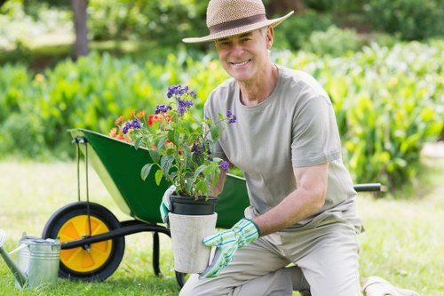 A mature woman is kneeling on a grassy lawn in a well-maintained garden, holding a potted flowering plant with purple and red blooms. She is wearing a light grey T-shirt, beige trousers, a wide-brimmed straw hat, and gardening gloves, smiling at the camera. Behind her, a bright yellow wheelbarrow with a green basin is positioned on the lawn, containing gardening tools and supplies. The garden features lush green grass, with a backdrop of tall hedges, shrubs, and mature trees providing shade and structure. The scene suggests a sunny day with natural light illuminating the vibrant colours of the plants and the outdoor environment, indicating active garden maintenance or planting work. This setting aligns with professional gardening services in the Sanderstead area, focusing on lawn care, planting, and outdoor landscaping.